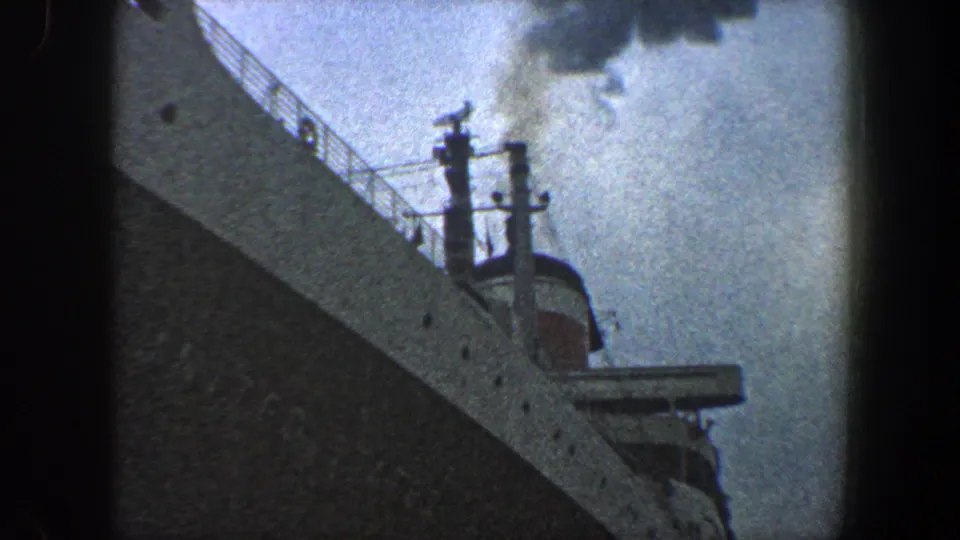 Low-angle 1960 New York City frame of the ocean liner's bow, railings, mast details, partial superstructure, and smoke against a pale cloudy sky.