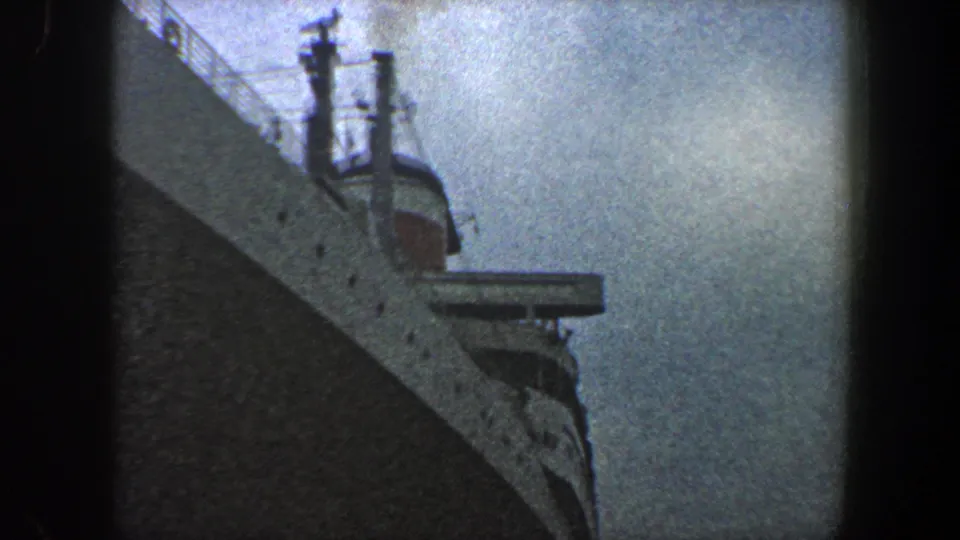 1960 New York City 8mm frame showing the ocean liner bow from below, with railings, a small projecting platform, and weathered sky filling the background.