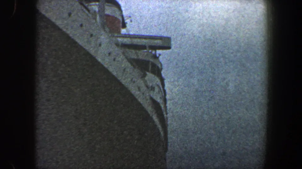 Final 1960 New York City frame with the ocean liner's bow dominating the lower view, upper deck structures visible near the top, and cloudy sky behind it.