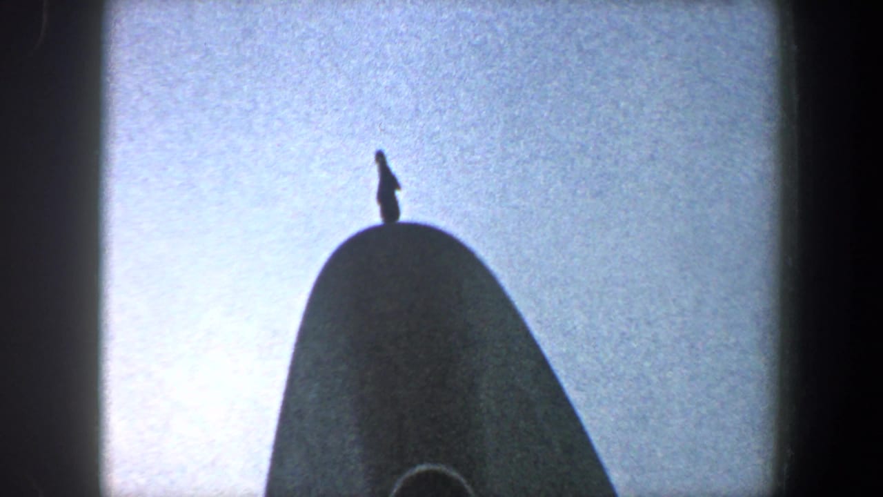 Small silhouetted person standing atop a curved ship structure against bright sky.