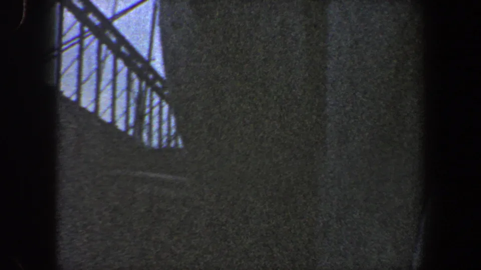 Grainy 1960 8mm frame of the ship hull at close range, with a railing at upper left and muted cloudy light.