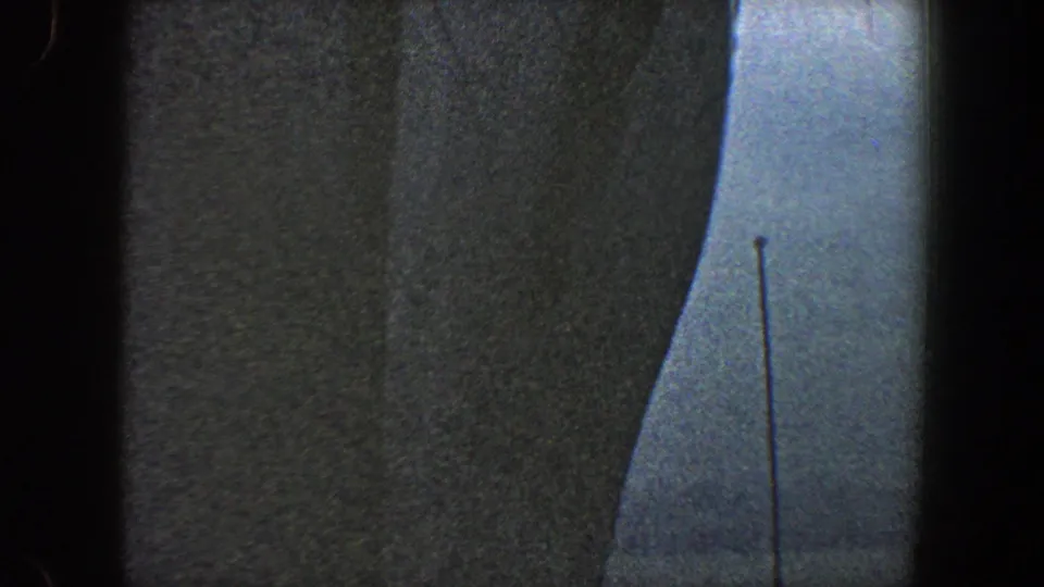 1960 New York City 8mm frame showing the curved edge of a large grey ship with pale sky and a thin vertical pole or mast nearby.