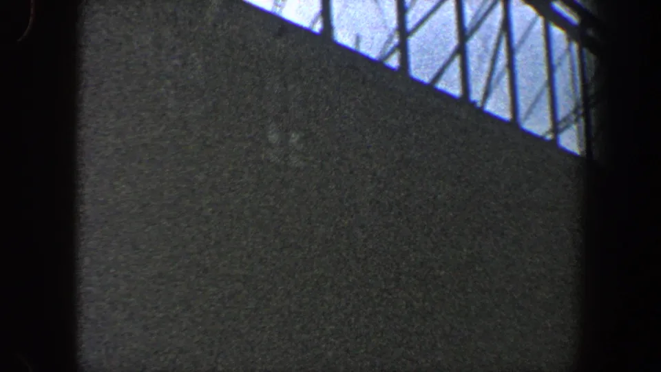 Grainy 1960 New York City 8mm frame looking up at a bridge under construction with diagonal steel framework visible overhead.