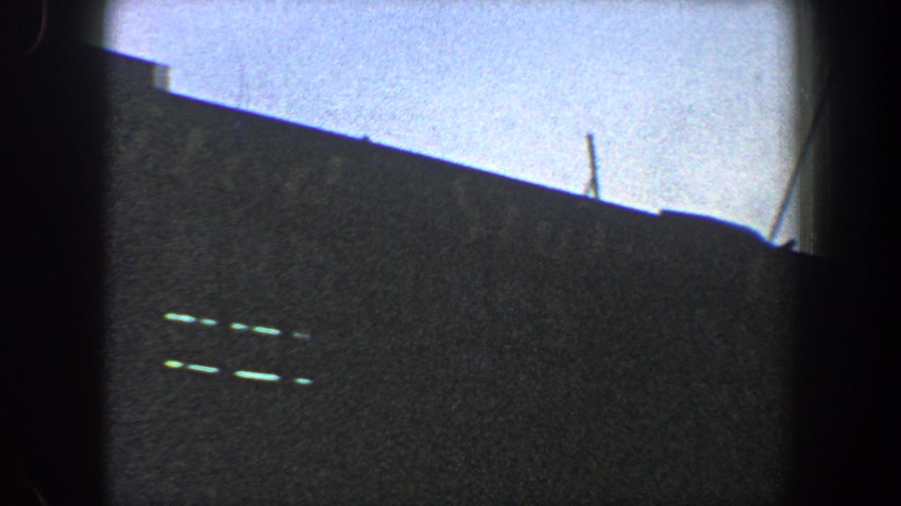 1960 New York City 8mm frame showing a dark building facade under pale blue sky, with faint horizontal details and visible film gate edges.