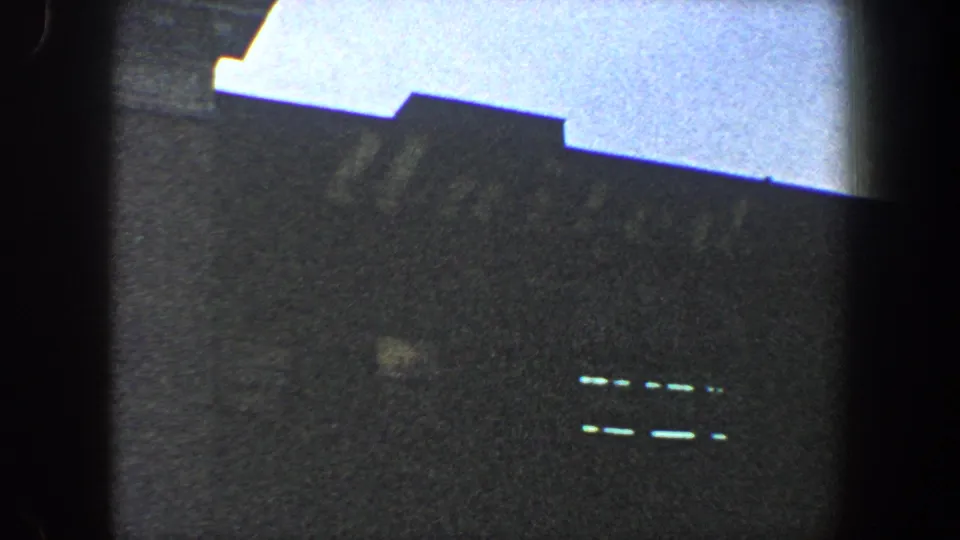 1960 New York City 8mm frame with a tilted roofline, dark facade, pale sky, and small bright horizontal markings on the building.