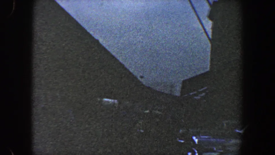 1960 New York City 8mm frame of a dark building corner beneath blue sky, with a slender pole or antenna near the right edge.