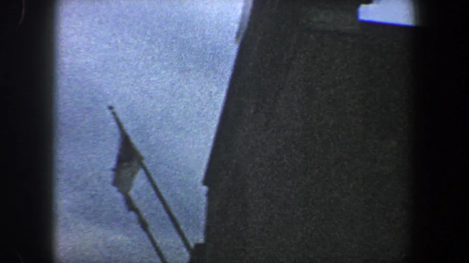 1960 New York City 8mm frame showing a tilted low-angle view of a flagpole, small flag, dark facade, and blue sky.