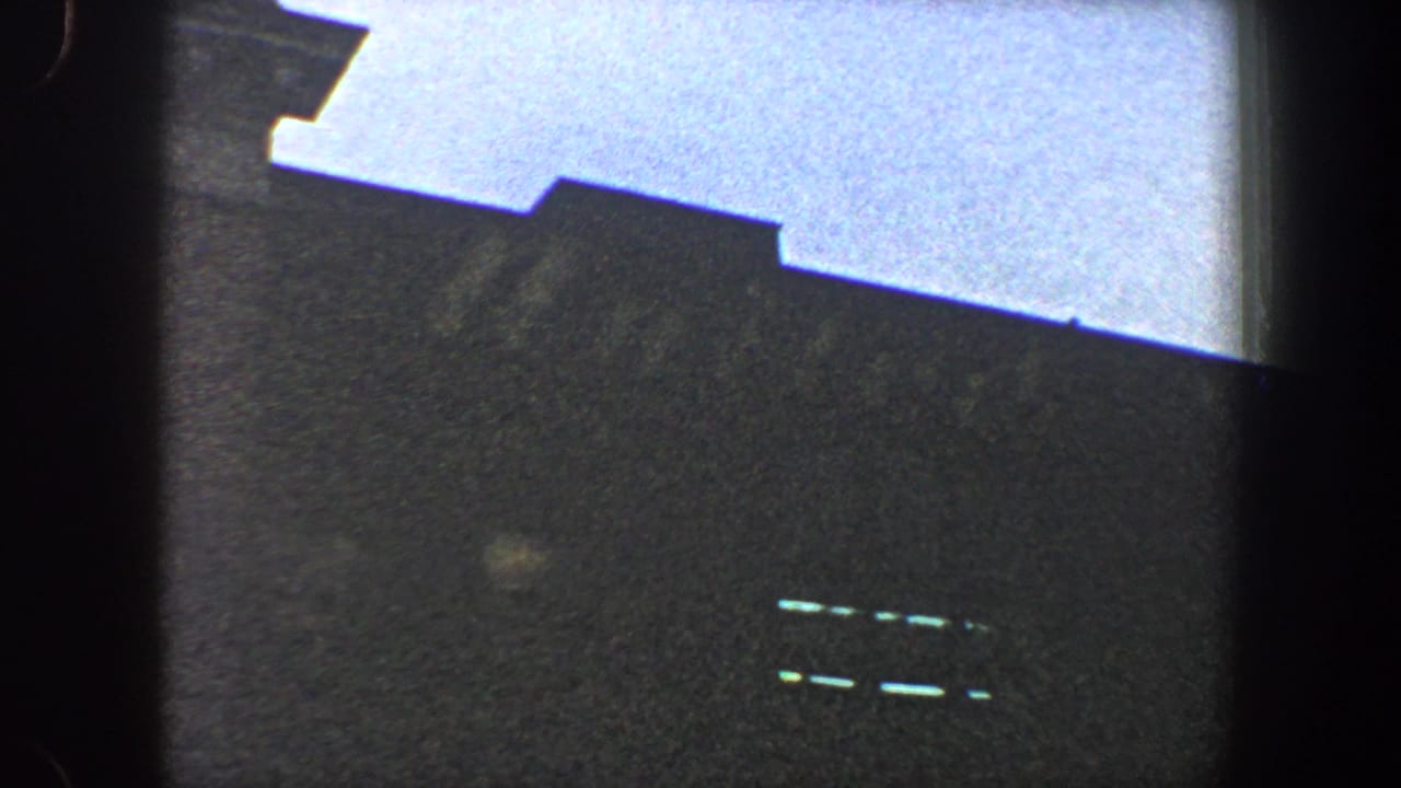 1960 New York City 8mm frame with a dark, poorly defined building or stadium form beneath a pale sky, with faint bright slits visible on the facade.