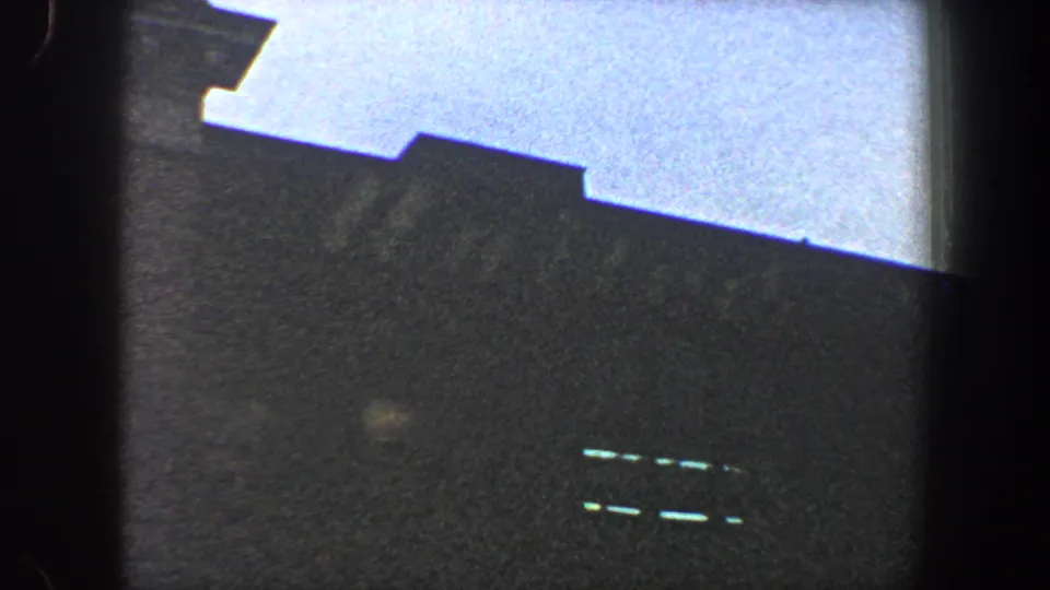 1960 New York City 8mm frame with a dark, poorly defined building or stadium form beneath a pale sky, with faint bright slits visible on the facade.