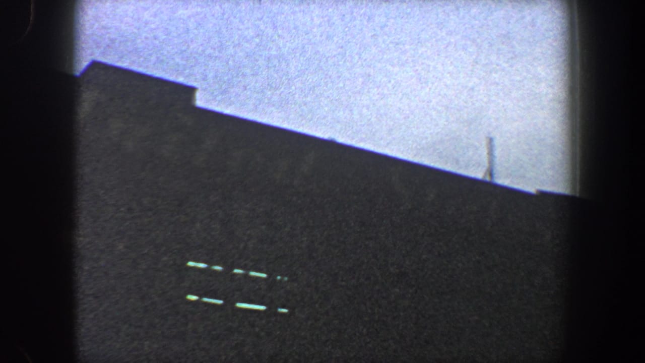 1960 New York City 8mm frame showing a sloping dark roof or stadium edge, faint bright openings, and a thin pole or antenna near the skyline.