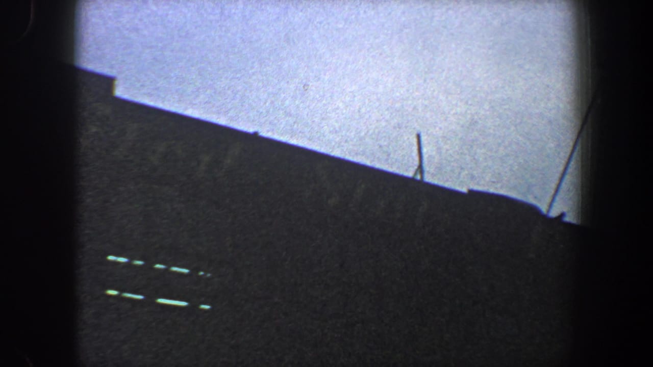 1960 New York City 8mm frame showing a dark sloped building or stadium edge with a small antenna or pole silhouetted against the pale sky.
