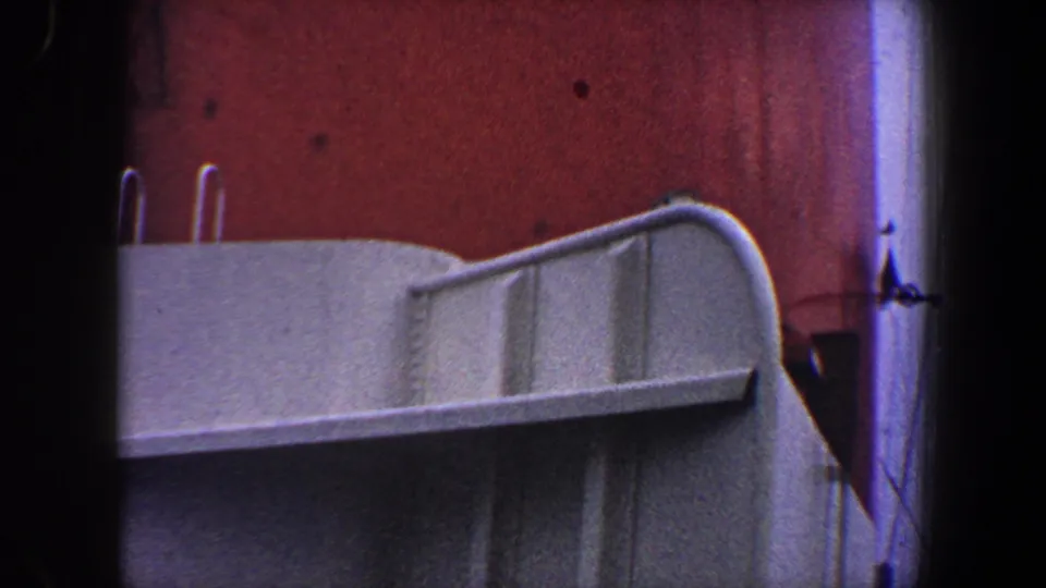 1960 New York City frame showing a white railing or deck edge crossing a red industrial wall, with dark frame edges from the 8mm scan.