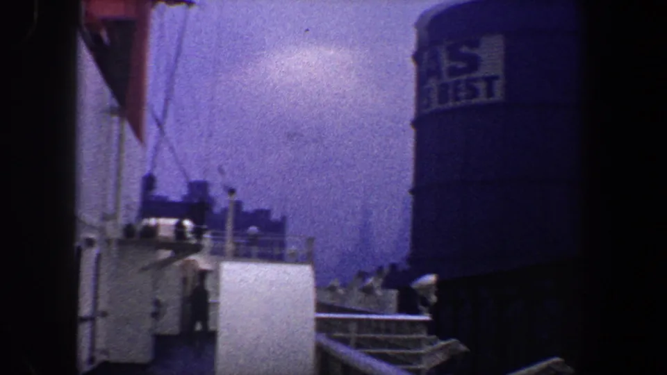 1960 New York City waterfront frame with ship structures, railings, possible distant people, and a blue tank with visible lettering.