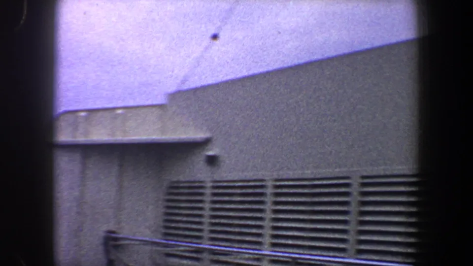 1960 New York City 8mm frame of a dockside or factory wall with long vents, railings, and hazy sky.
