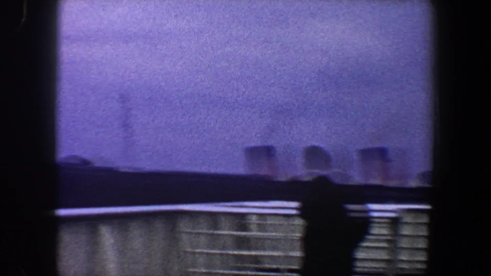 A silhouetted figure passes near the railing on a New York City boat deck, with hazy waterfront structures in the background.