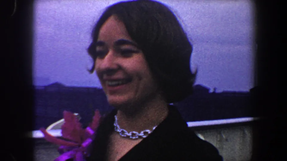 A smiling woman in a dark jacket appears close to the camera on a 1960 New York City boat, with a bright pinned flower visible.