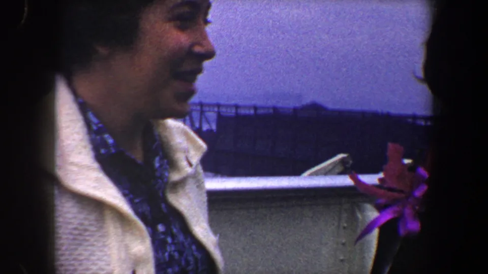 A woman in a light coat smiles in profile on a 1960 New York City boat deck, with another person and a bright flower at the edge.