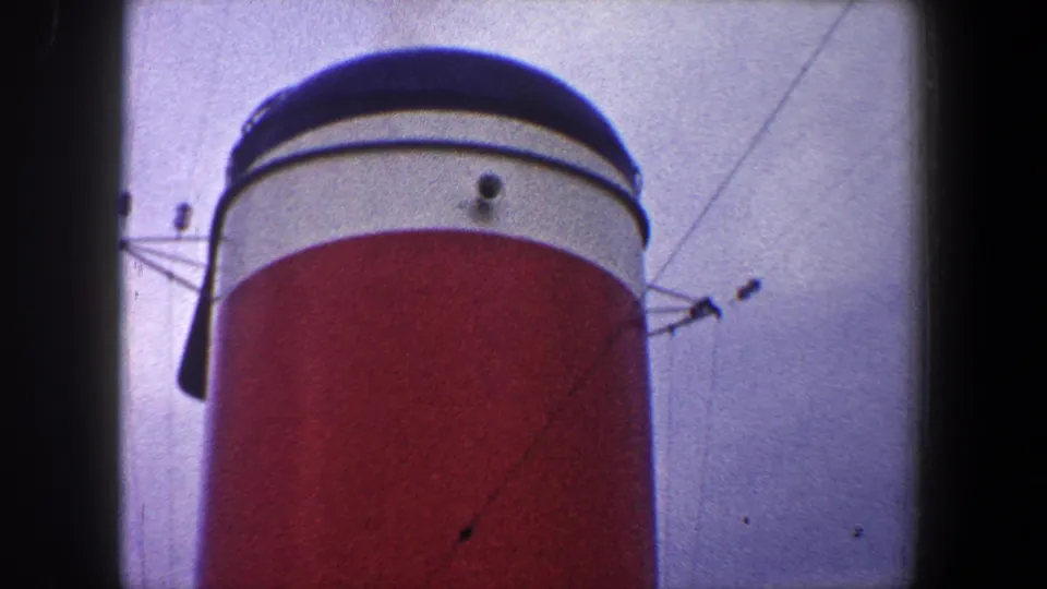 A red cylindrical smokestack with a dark cap fills the frame in grainy 8mm footage from the New York City clip.