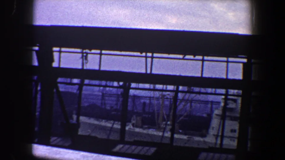 1960 New York City home-movie view through a dark ship railing toward water and a nearby vessel under bright sky.