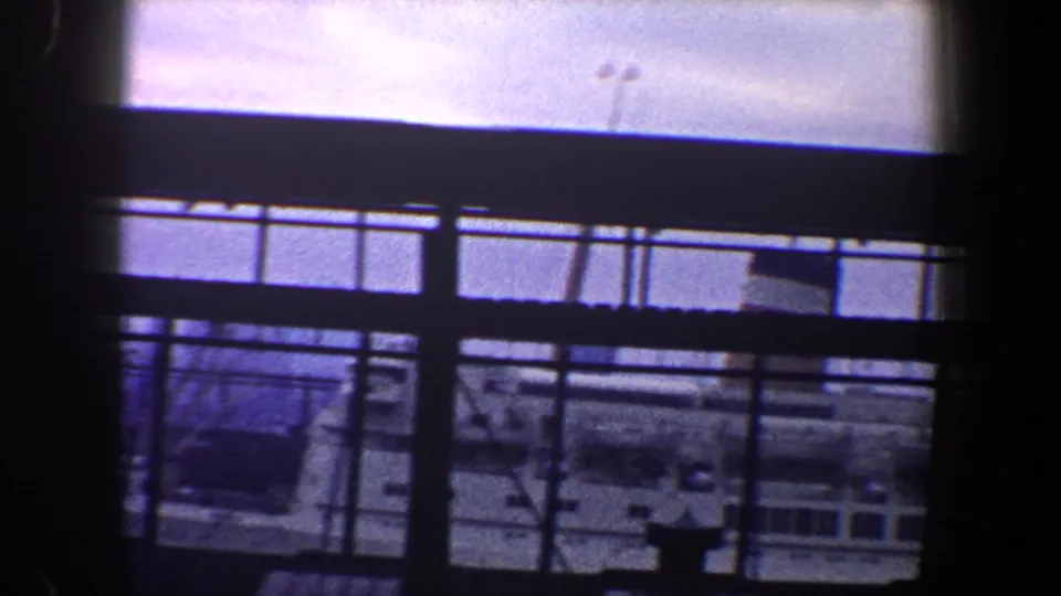 A handheld 8mm view from a ship deck in New York City, with dark railing bars cutting across water and a nearby vessel.
