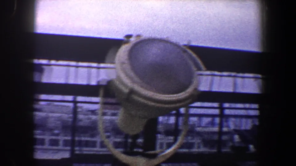 Close shipboard detail with a round deck fixture silhouetted against railings, water, and a nearby vessel in 1960 New York City footage.