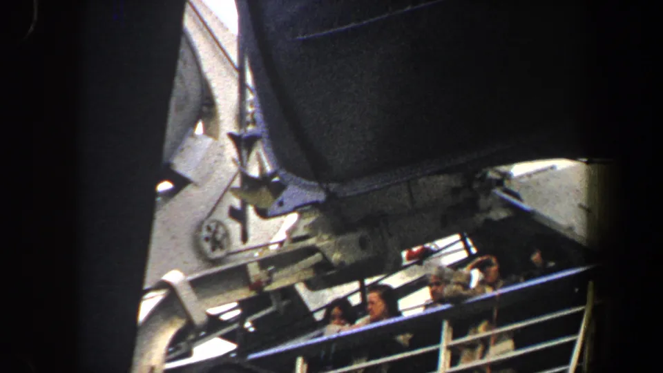 Handheld 8mm view of passengers behind a ship railing, partially blocked by large vessel structure during departure.