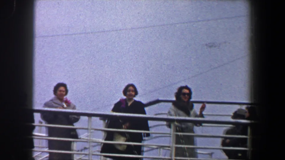 1960 New York City departure frame with women in coats standing at a ship railing and one passenger waving toward the dock.