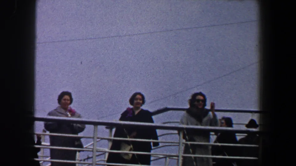 1960 New York City home-movie frame of three women in winter coats standing at a ship railing and looking toward the camera.