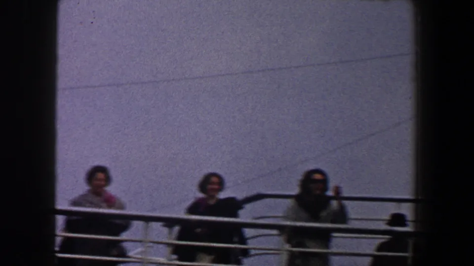 1960 New York City ship-deck frame with three women along a white guardrail, one raising a hand toward the camera.