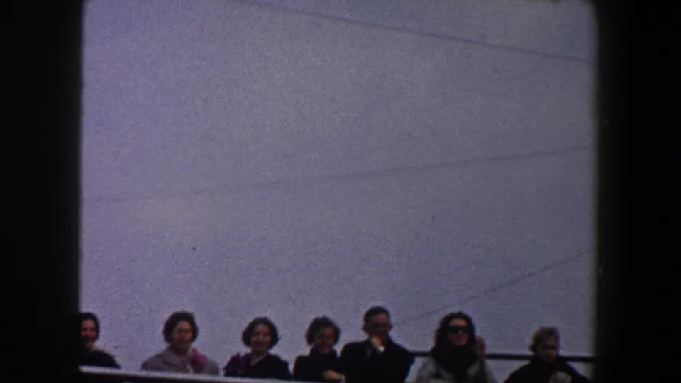 Adults in coats stand behind a ship railing in 1960 New York City home-movie footage, with a dark vertical frame at left.