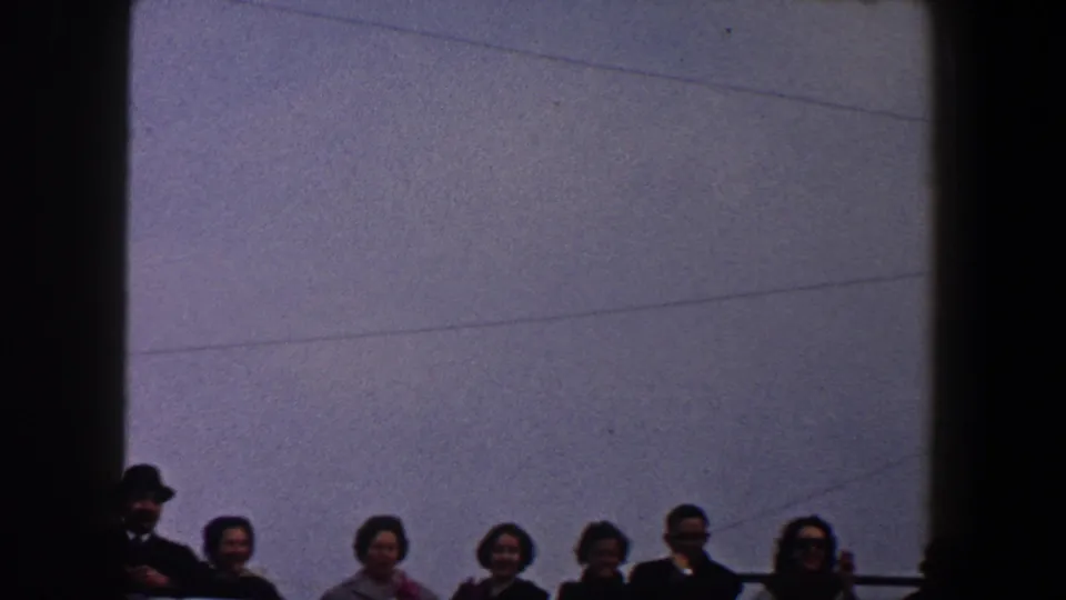 A group of adults stands at a railing in 1960 New York City footage, with coats, sunglasses, and a man wearing a dark brimmed hat.