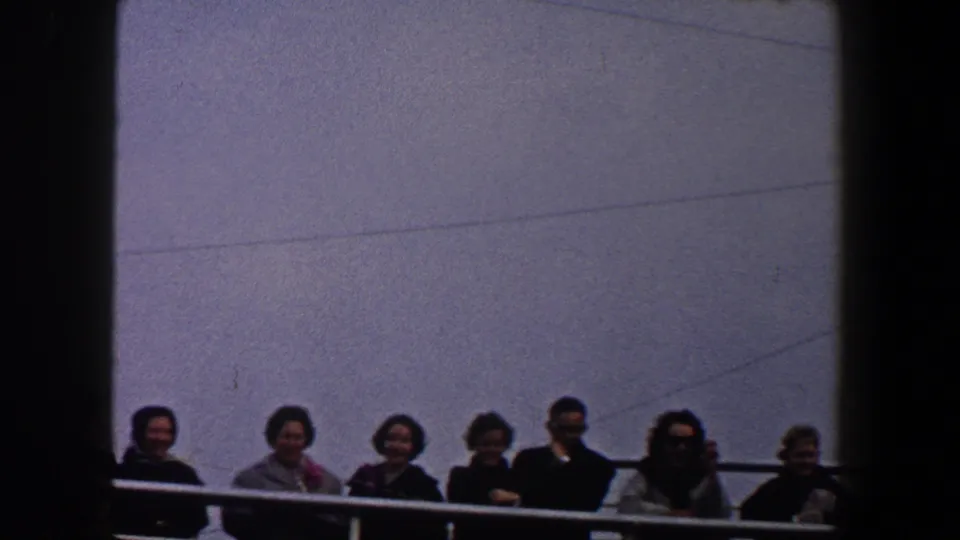 Adults in mid-century clothing stand behind a railing in 1960 New York City home-movie footage, framed by dark vertical divisions.