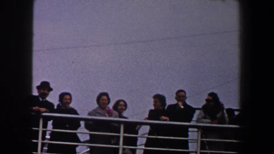 Adults in coats stand and lean at a ship railing in 1960 New York City footage, some looking outward and others turned toward one another.