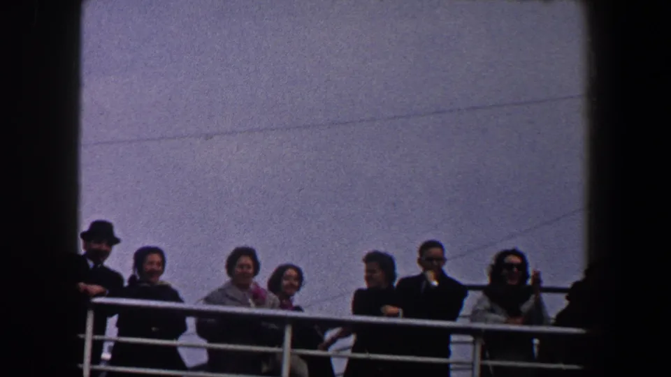 1960 New York City home-movie frame showing adults in coats, suits, and hats along a vessel railing under open sky.