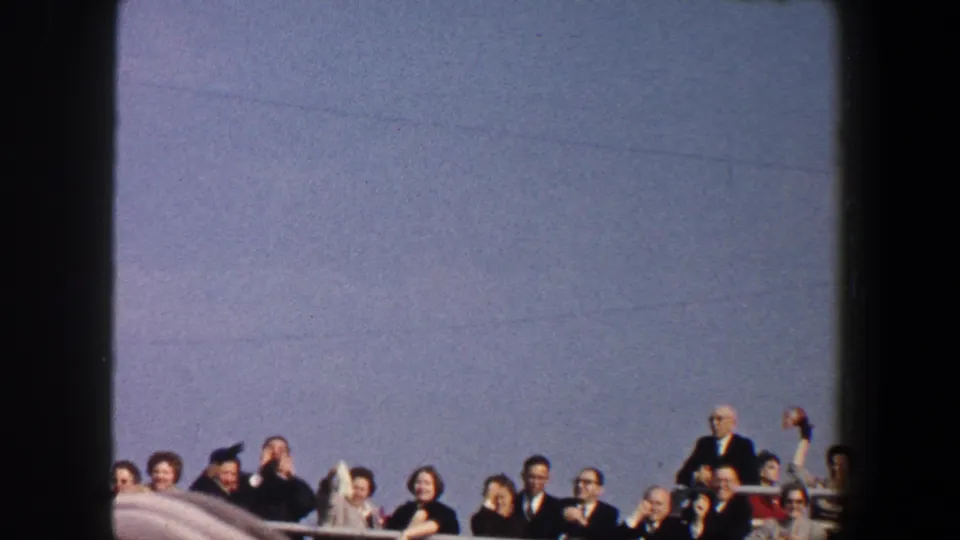 1960 New York City frame of a boat or ferry crowd waving and smiling from behind a railing beneath a wide blue sky.