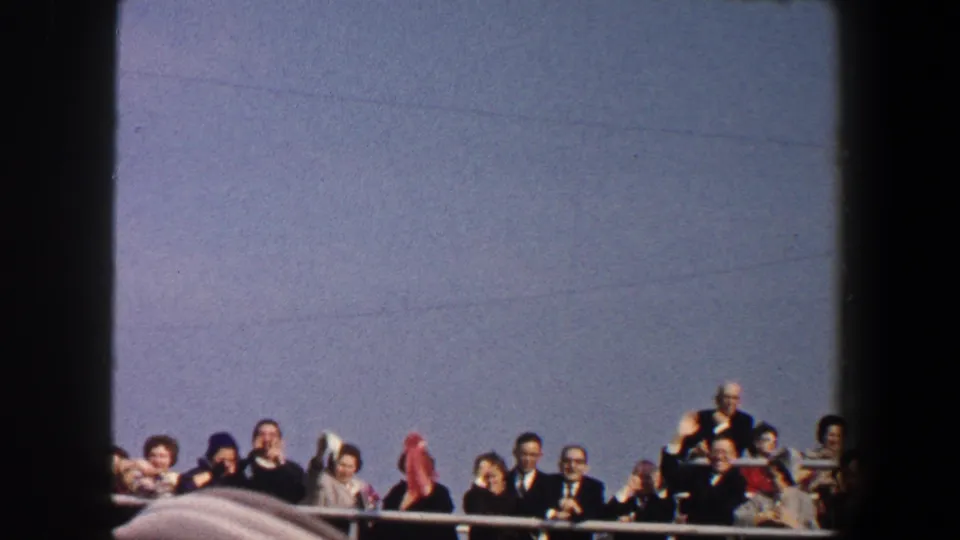 1960 New York City frame of well-dressed adults waving from a boat or ferry railing, with faded color and visible film borders.