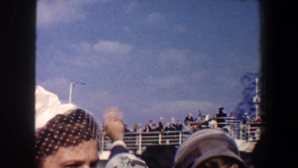 1960 New York City 8mm frame showing foreground passengers and a distant deck crowd waving across the vessel.