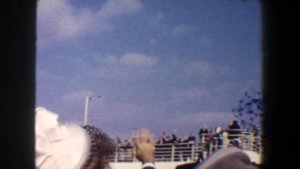 1960 New York City 8mm frame with foreground hats, a raised hand, and passengers waving from a boat deck.