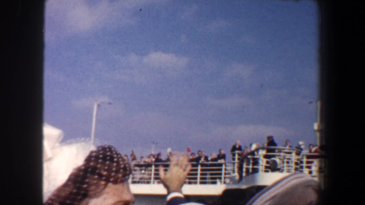 Crowd on a ship deck under blue sky, with a woman in a netted hat waving in the foreground.