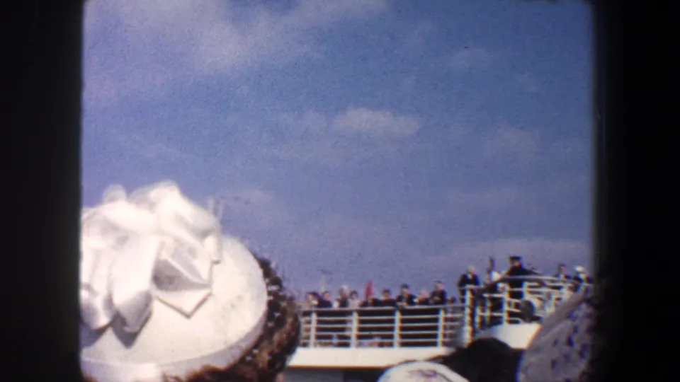 Foreground woman in a white decorated hat watches passengers gathered along a ship deck during a 1960 New York departure.