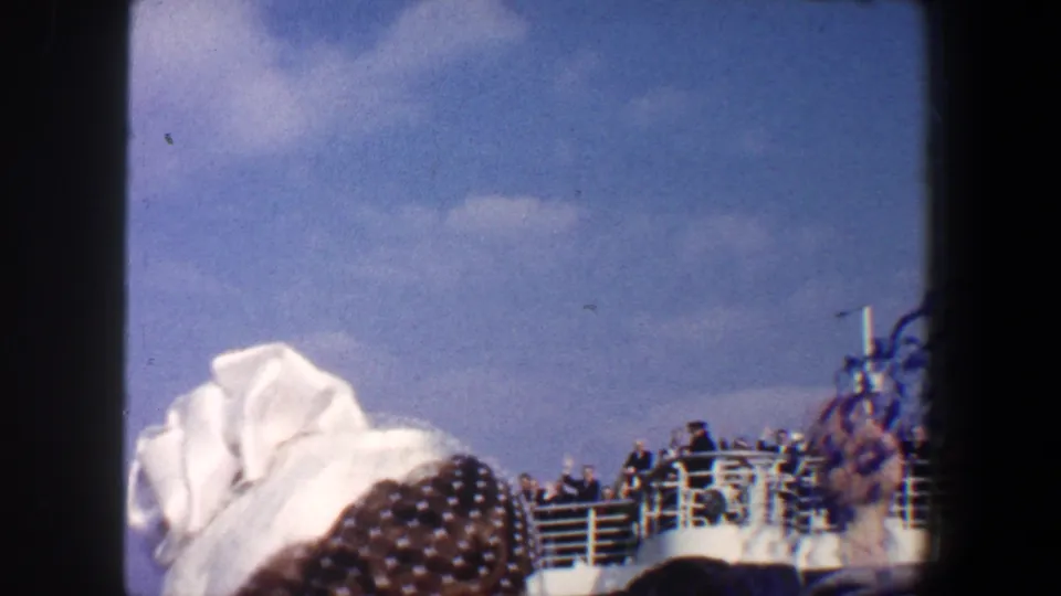 A woman in a white hat and veil is seen from behind while passengers line the deck of a departing ship in New York City.