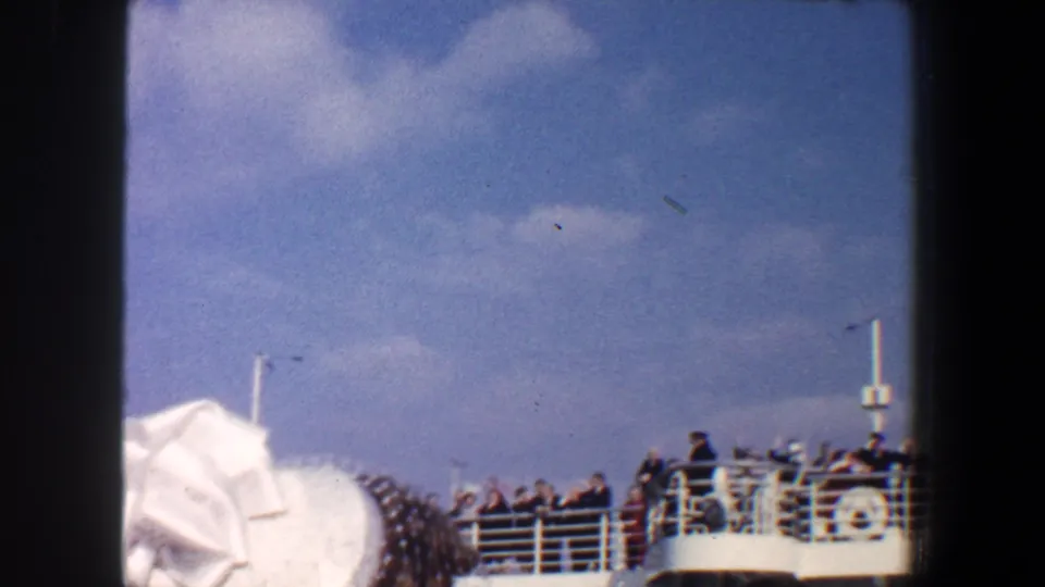 Passengers stand along a ship deck railing under blue sky, seen over foreground hats during a 1960 New York City farewell.