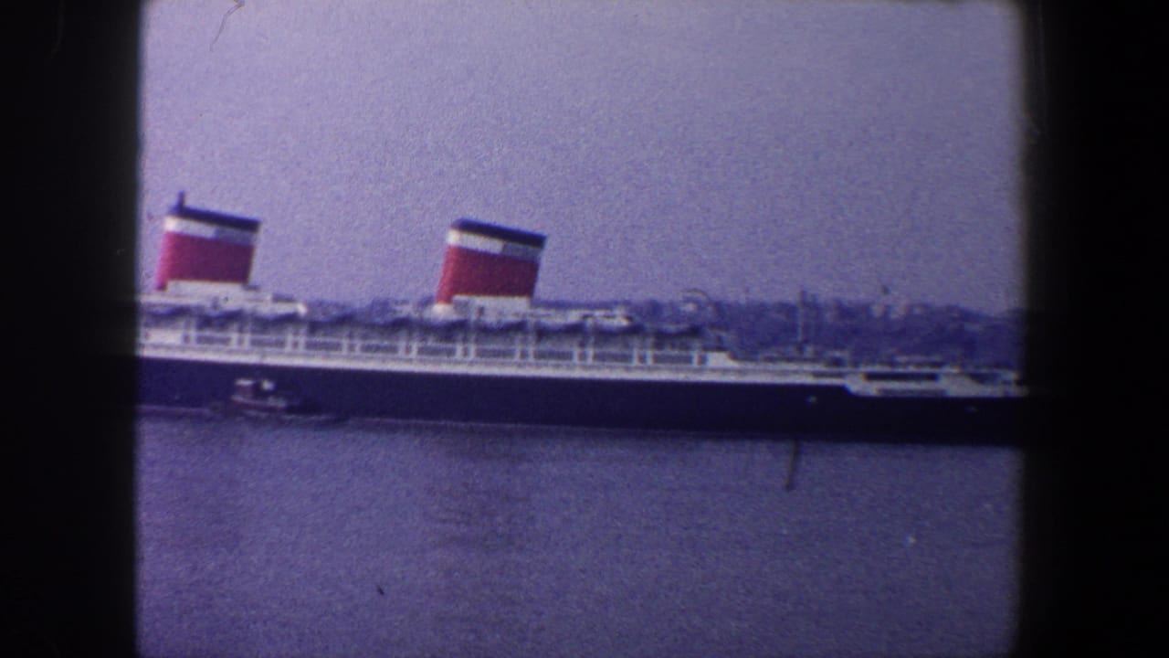 1960 New York City home-movie frame showing a large ocean liner docked across the water with red funnels and hazy shoreline behind it.