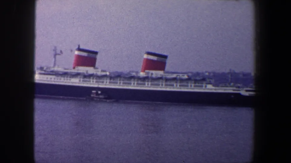 1960 New York City harbor view of the docked ocean liner, with a small tugboat near the dark hull and water in the foreground.