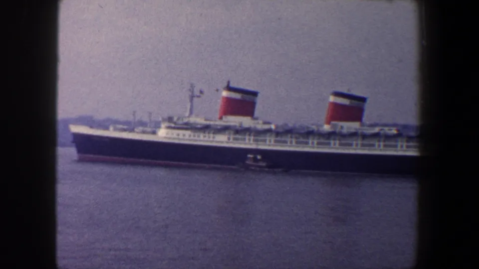 1960 home-movie frame of a two-funnel ocean liner in New York harbor, filmed in soft color from a distance.