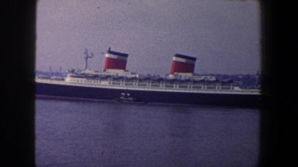 Large ocean liner in New York harbor, partly cropped at the right edge, with tugboat and distant shoreline visible.