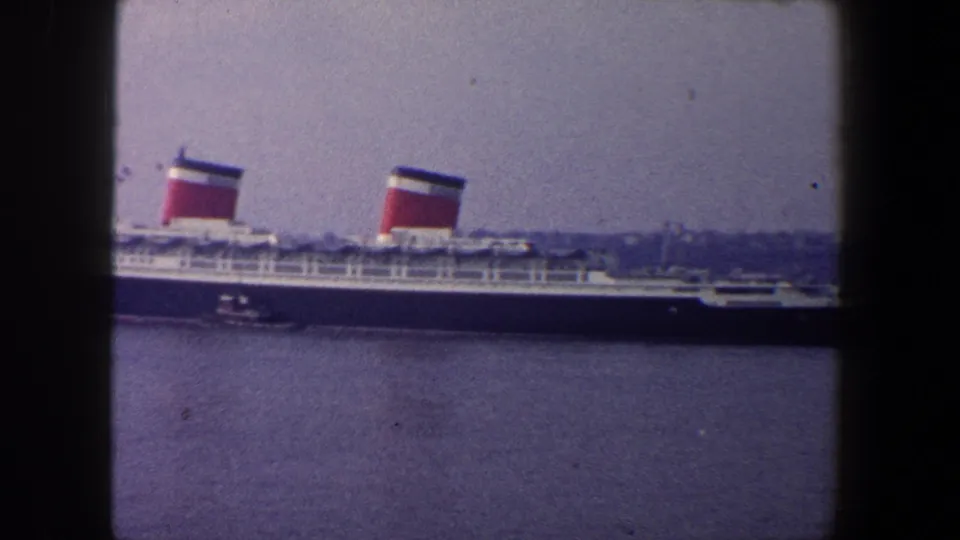 Large ocean liner in 1960 New York City harbor, seen broadside with two funnels and a small tugboat near the hull.