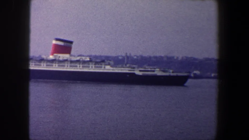 Partial view of the ocean liner moving through New York City harbor, with one funnel prominent and shoreline in the distance.