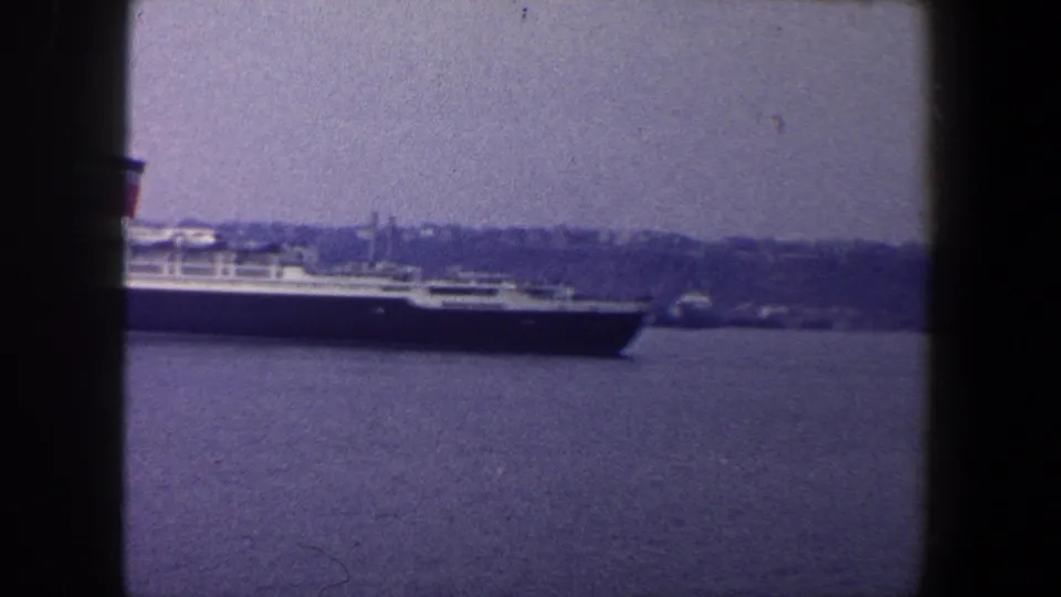 End of the 1960 harbor shot as the ocean liner passes partly out of frame, leaving open water and distant shoreline visible.