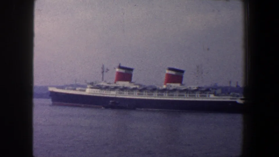 1960 New York City 8mm frame showing a large passenger liner passing on calm water with a hazy shoreline beyond.